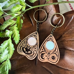 May include: Two wooden keychains shaped like a witch's  ouija board with a  circular iridescent  charm in the centre.  One charm is white and the other is blue.
