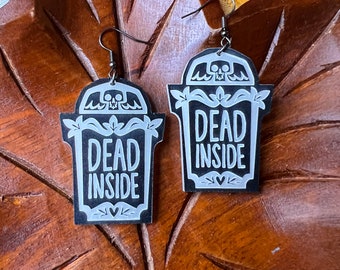 May include: A pair of black and silver earrings shaped like tombstones with the text "Dead Inside" on each. The earrings have a skull and crossbones design at the top.