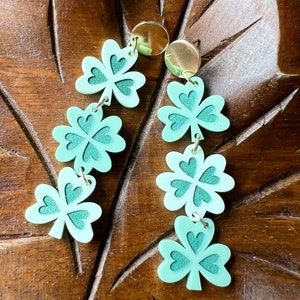 May include: A pair of dangle earrings featuring four green shamrock charms with gold accents.