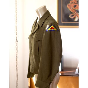 May include: Olive green military jacket with a colorful patch on the shoulder. The jacket has a collared neckline, two chest pockets, and buttoned cuffs. The patch features a yellow, blue, and red design. The jacket is displayed on a mannequin.