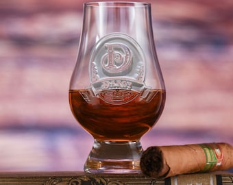 May include: A clear glass whiskey tumbler with a personalized engraving of the letter "D" and the name "Darcy". The tumbler is filled with amber-colored whiskey and is sitting on a book with a cigar.