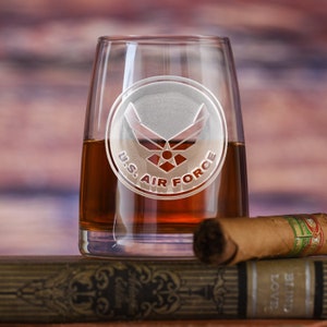 May include: A clear glass tumbler with an etched design of the U.S. Air Force logo. The glass is filled with a dark amber liquid and is sitting on a stack of books.