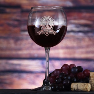 May include: A personalized red wine glass with a shield design and the letter "R" engraved on it. The shield has a banner with the name "Reese" on it. The glass is filled with red wine and is sitting on a wooden surface with grapes and a cork.