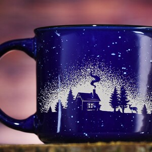 May include: A blue ceramic mug with a white design of a cabin in the woods with a snowy background. The mug has a speckled blue glaze.