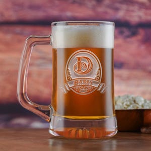 May include: A clear glass beer stein with a handle. The stein is filled with a light amber beer. The stein is engraved with a monogram and the name "Darcy".