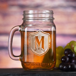 May include: A clear glass mason jar mug with a handle, etched with a crest and the letter "M". The mug is filled with a light brown liquid and is sitting on a dark surface next to a bunch of green and purple grapes.