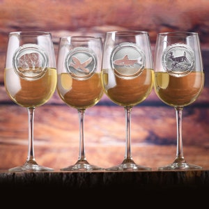 May include: Four clear wine glasses filled with white wine. Each glass has a decorative metal charm with an animal design: a bear, a bird, a shark, and a deer. The glasses are arranged on a wooden surface.