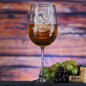 May include: A clear glass wine glass with an etched design of Italy and the text "Antonini" on the side. The glass is filled with white wine and is sitting on a wooden surface with grapes and a wine cork.