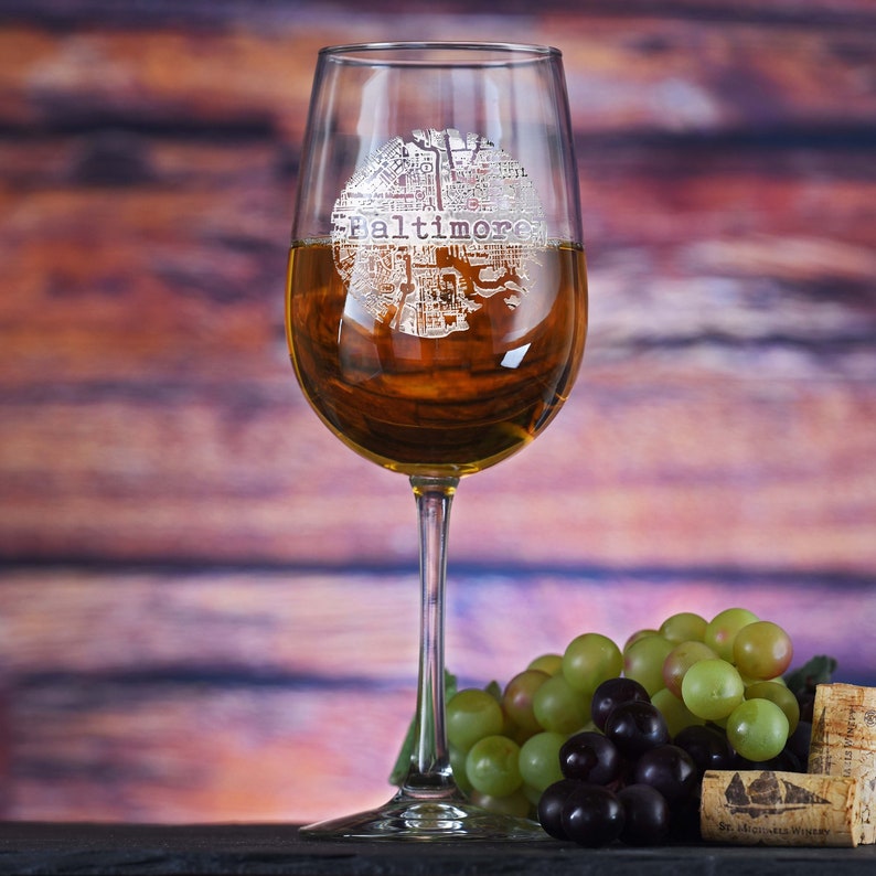 May include: A clear wine glass with a design etched into the side. The design is a map of Baltimore, Maryland. The glass is filled with white wine and is sitting on a wooden surface. There are grapes in the background.