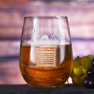 May include: A stemless wine glass filled with amber liquid. The glass is etched with an eagle and a scroll bearing text from the 2nd Amendment. Grapes are visible in the background. The glass is on a dark surface.