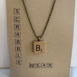 May include: A brown card with the word "SCRABBLE WEAR" printed on it. A gold chain necklace with a wooden Scrabble tile pendant that says "B3" is hanging from the card.