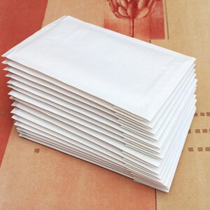 Op de afbeelding: Een stapel van twaalf rechthoekige witte papieren enveloppen met verzegelde randen. De enveloppen zijn netjes in een verticale stapel geplaatst, met een gladde, matte afwerking en uniforme afmetingen voor een strak uiterlijk.