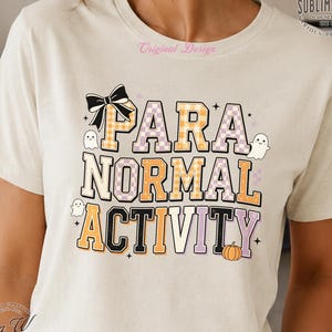 Puede incluir: Una camiseta de color crema con el texto "PARA NORMAL ACTIVITY" en una fuente de cuadros. El diseño incluye un lazo negro, tres pequeños fantasmas blancos y una pequeña calabaza naranja, decorado con diminutas estrellas negras en el frente.