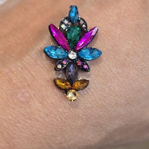 May include: A multi-colored jewel-encrusted floral design with a black base. The design features blue, green, pink, purple, and yellow gemstones. The image includes the text "Christine Beaudry".