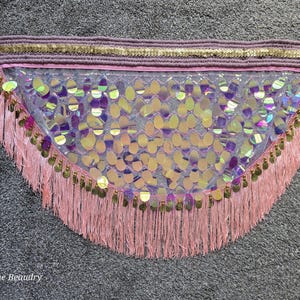 May include: A belly dance hip scarf with iridescent sequins, pink fringe, and gold accents. The top band is lavender with gold trim, and the ties are lavender. The design is semi-circular.