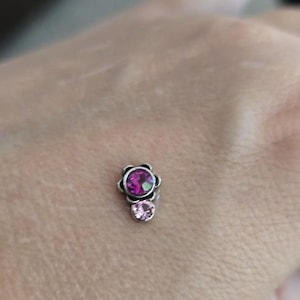 May include: A small, silver-toned floral stud earring with a large, round, magenta-colored gemstone at the top and a smaller, round, light pink gemstone below. The earring is designed with a flower-shaped setting.