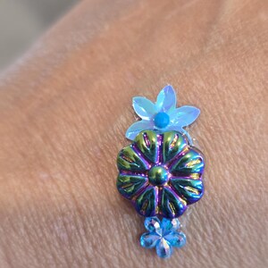 May include: A floral-shaped embellishment with iridescent green, blue, and purple hues. The design features three flowers of varying sizes, with a small blue bead at the center of the top flower. The embellishment is likely for crafts or jewelry.