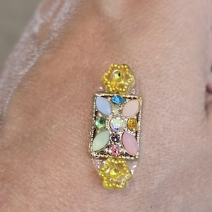 May include: A gold-toned rectangular brooch with a floral design, featuring a central array of colorful gemstones in pastel hues. The brooch has two yellow flower accents and the text "Christine Beaudry".