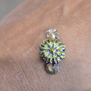 May include: A small, decorative flower-shaped embellishment with a gold center and blue-edged petals. Two clear, faceted crystals are positioned above and below the flower. The object is likely a jewelry component or decorative element.