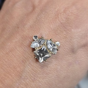 May include: A gold-toned brooch featuring a central square-cut clear crystal surrounded by smaller, marquise-shaped and round crystals. The brooch has a cluster design, with the crystals set on a gold-colored metal base.