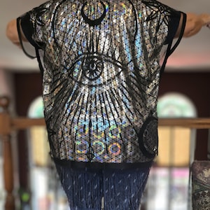 May include: Black mesh kimono with a silver holographic design featuring an eye with a crescent moon on either side. The kimono has a fringe trim.