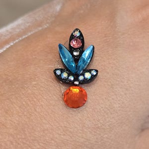 May include: A decorative face jewel with a black base, featuring blue and pink crystals in a floral design, and a large orange crystal at the base. The jewel is designed to be applied to the face or body.