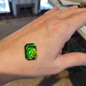 May include: A green and yellow iridescent gemstone with a black border. The gemstone is rectangular in shape and has a smooth, polished surface.