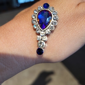 May include: A silver teardrop-shaped pendant with a large blue gemstone surrounded by smaller clear gemstones. The pendant is attached to a silver chain with a smaller blue gemstone at the end.