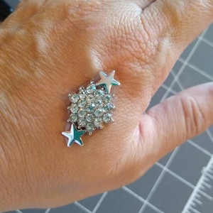 May include: A silver-toned ring with a cluster of small, clear crystals and two teal star accents. The ring is set against a neutral background, showcasing its sparkling details. The ring is a piece of jewelry.