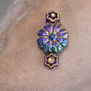 May include: A close-up of a decorative floral bead with iridescent purple, blue, and gold hues. The bead has a large flower shape in the center, with smaller flower shapes above and below. The bead is set against a neutral background.
