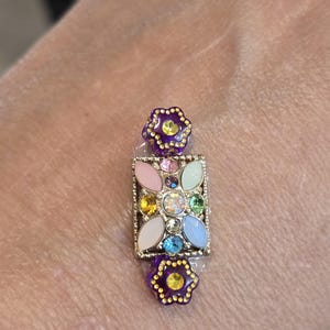 May include: A decorative, rectangular brooch with a gold-colored frame. The brooch features a central design of colorful gemstones and is flanked by two purple flower-shaped accents. The brooch is set against a skin-toned background.