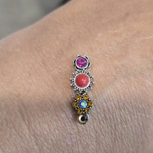 May include: A silver-toned charm with three decorative elements. The top features a pink gemstone in a floral setting, followed by a red gemstone in a sunburst design, and a yellow and blue gemstone flower at the bottom. The charm is likely for a bracelet or necklace.
