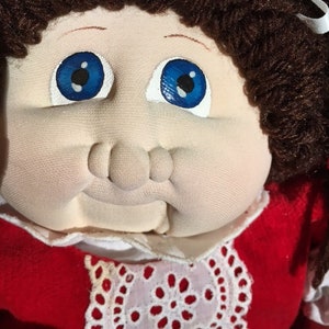 May include: A Cabbage Patch Kid doll with brown yarn hair, blue eyes, and a red dress with white lace trim.