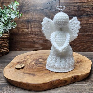 May include: A white crochet angel figurine with a halo and wings, displayed on a wooden slab. The angel is handmade. The background includes a wooden wall and a small potted plant.