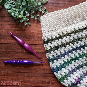 May include: A close-up of a crocheted hat with a white base and green, blue, and purple stripes. Two purple crochet hooks are lying on a wooden surface.