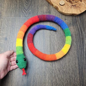 Op de afbeelding: Een gehaakt slangen-speeltje met een levendig regenboogpatroon in segmenten van rood, oranje, geel, groen, blauw en paars. De slang heeft twee zwarte plastic ogen en een kleine rode gehaakte tong die uit de groene kop steekt.
