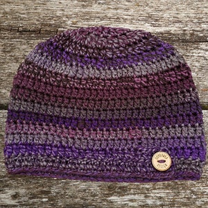 Puede incluir: Una mujer con una chaqueta vaquera, una camisa negra y un gorro y una bufanda de ganchillo morado y gris. El gorro tiene un botón marrón en la parte superior. La mujer está de pie en un bosque con follaje otoñal.