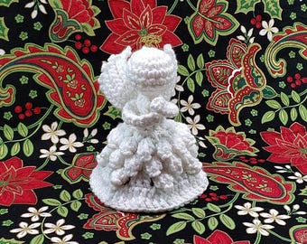 May include: White crocheted angel ornament with a ruffled skirt and wings. The angel is standing on a red and green floral patterned fabric.