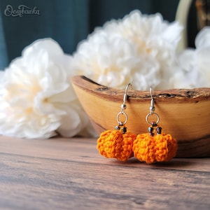 May include: A pair of orange crocheted pumpkin earrings with silver hooks and beads. The earrings are displayed on a wooden surface with white flowers in the background.