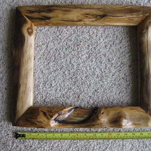 May include: A rectangular wooden picture frame with a natural live edge and visible wood grain patterns in shades of brown and tan. A yellow measuring tape is positioned along the bottom edge, showing a length of approximately 20 inches.