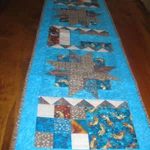 May include: A rectangular quilted table runner with a light blue background and a variety of patterned fabric squares. The design includes star shapes, arrows, and deer in shades of brown, gray, and blue. The runner is bordered with a gray fabric.