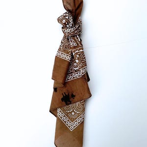 May include: A brown bandana featuring a white paisley pattern, tied into a knot at the top. The fabric displays a distinct brown colour with white decorative motifs and a dark, irregular smudge mark visible on the lower section of the folded cloth.