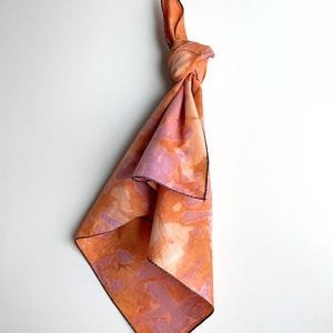 May include: A knotted, square scarf with a tie-dye pattern in shades of orange, pink, and white. The edges are finished with a black stitch. The scarf is hanging against a white background.