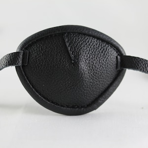 Könnte beinhalten: Ein schwarzes Leder-Augenpflaster mit verstellbarem Band. Das Pflaster ist so konzipiert, dass es ein Auge bedeckt.