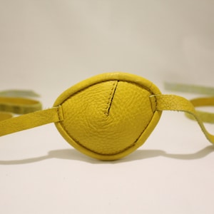 Könnte beinhalten: Ein gelbes Leder-Augenpflaster mit einem Band. Das Augenpflaster ist aus strukturiertem Leder und hat ein schlichtes Design.