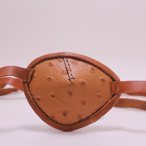 May include: A brown leather eye patch with a textured surface and a strap.