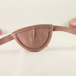 Op de afbeelding: Een roze leren ooglapje met een witte gestikte rand en een witte gestikte band.