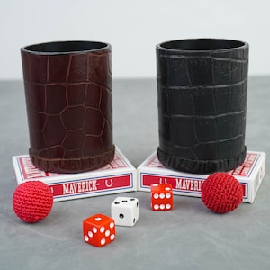 Può includere: Due bicchieri da dado in pelle, uno marrone e uno nero, sono appoggiati su due mazzi di carte da gioco. Le carte sono etichettate "Maverick". Ci sono quattro dadi e due palline rosse sul tavolo.