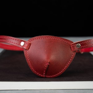 May include: A burgundy leather eye patch with a matching strap and red interior. The eye patch is stitched and has silver-colored rivets. The strap is designed to go around the head.