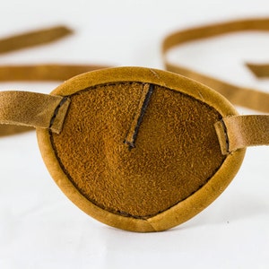 May include: A brown leather eye patch with a strap. The patch is made of suede and has a stitched edge.